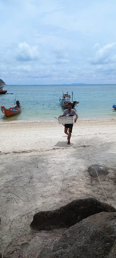 A man carries a white object on the beach in Baan Chaloklum, Thailand. The man, wearing a light-colored patterned shirt and dark shorts, is on the sandy beach with several fishing boats in the turquoise water. A large rock formation is visible to the right. The sky is partly cloudy.