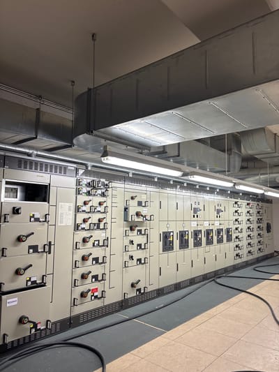 The image shows a room containing a large electrical distribution panel. The panel is composed of multiple modular sections, each with various switches, levers, and indicator lights. Large metallic ductwork is visible overhead, running across the ceiling. Black electrical cables are present on the floor, some coiled and others extending across the room. The walls are a neutral color, and the floor appears to be tiled. There are no people visible in the image. This appears to be a utility room or an electrical substation within a building in the city of Mannheim, Germany.