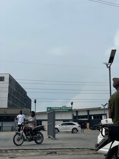 In the city of Ojomu, Nigeria, a man is riding a red motorcycle with a passenger on the back. The passenger is wearing a patterned long-sleeved shirt and khaki shorts. The motorcyclist is wearing a white t-shirt and dark pants. The scene appears to be in front of a supermarket called "PRINCE EBANO SUPERMARKET." Two other individuals are visible in the background, one standing by a fence and another walking near the supermarket entrance. Several cars are parked in the vicinity. The sky is overcast.