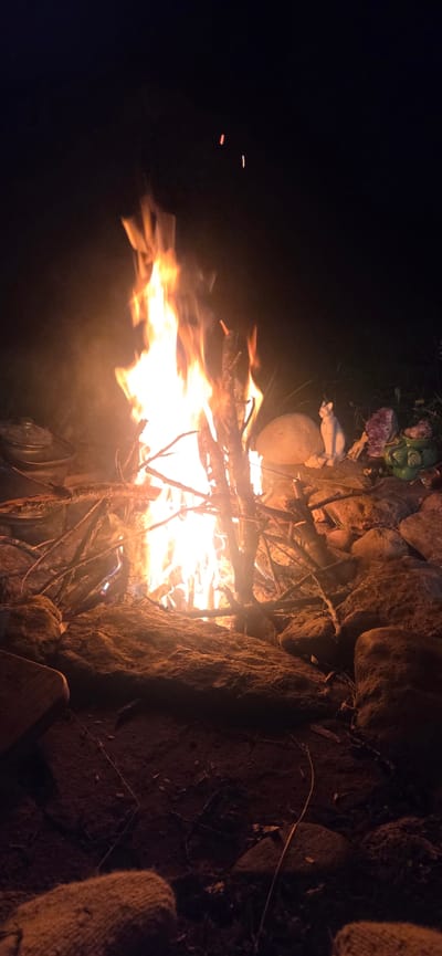A campfire burns brightly at night, illuminating a pile of sticks and rocks. In the background, there are several decorative objects, including a white cat statue, a purple crystal, and a green ceramic pot. The scene appears to be outdoors, possibly in a natural setting. The location is not specified as being in the city of Lesidren, Bulgaria.