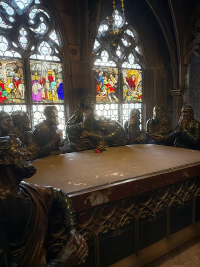 The image depicts a table surrounded by eleven bronze sculptures of men, seemingly engaged in a discussion or meal. The sculptures are arranged around a large, stone table. Behind the table are three Gothic stained-glass windows, depicting various religious scenes in vibrant colors. Above the central window, a large, ornate chandelier hangs from the ceiling. The overall setting appears to be an interior space with dark, carved wooden paneling and stone architectural elements, suggestive of a historical building. This scene is located in the Freiburg Minster in Freiburg im Breisgau, Germany, and the sculptures are part of a depiction of the Last Supper.