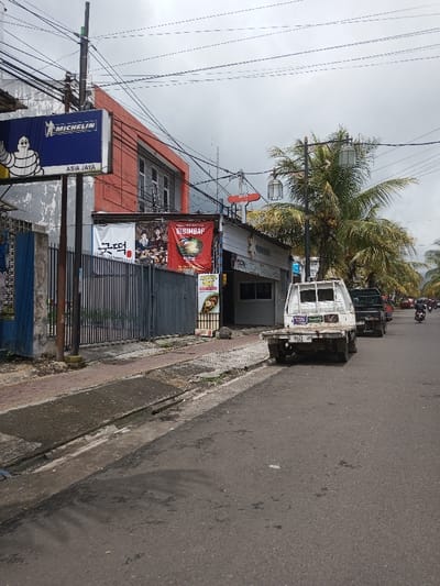 This image depicts a street scene in the city of Vaatharawattai, Sri Lanka, under an overcast sky. The view is angled towards the left side of a paved asphalt road, revealing a narrow, uneven sidewalk bordered by a curb and a drainage channel.
On the left, a blue billboard featuring the Michelin Man and the text "MICHELIN" with "ASIA JAYA" below is prominent. Behind this sign, a two-story building with a red upper facade and a grey lower section with windows is visible. Adjacent to it, a grey metal fence or gate precedes a single-story building. This building displays a white banner with Korean text and images of people and food, alongside a red banner advertising "BIBIMBAP" with a food illustration. A smaller, vertical yellow and red banner with "HAPPY DAYS" and a food item is also present. Numerous overhead power lines and utility poles traverse the scene.
On the asphalt road, a white flatbed truck is parked, facing away from the viewer. Its license plate "F 9761 JP" is visible on the rear, along with several stickers on the back window. Behind it, a dark-colored pickup truck or SUV is partially observed. Further down the road, on the right side, multiple motorcycles with riders are in motion.
To the right of the parked vehicles and sidewalk, large palm trees, some bearing coconuts, are visible. An ornate black streetlight pole with two lanterns is positioned in front of these trees. A small red satellite dish is mounted high on a pole in the background. No identifiable individuals are present in the immediate foreground or midground.