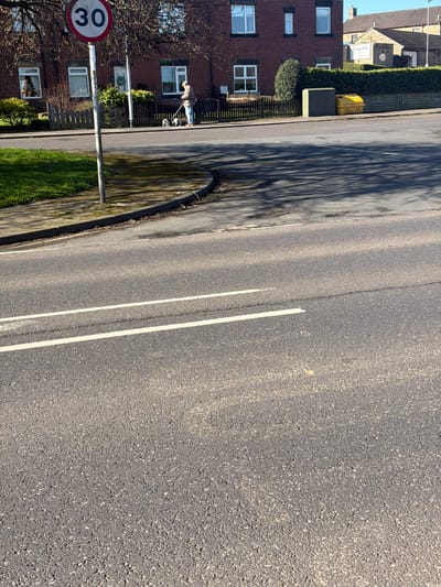 The scene depicts a residential street in Rotherham, United Kingdom, under clear, sunny daytime conditions.
In the foreground, an asphalt road surface occupies the lower half of the frame, marked with two parallel solid white lines indicative of a lane division. Shadows, consistent with tree foliage, are cast across the road surface. A concrete curb delineates the road from an elevated grassy verge on the left.
In the mid-ground, a grey metal pole displaying "S2950" vertically supports a circular red-bordered speed limit sign with "30" (miles per hour) printed in black. This pole is situated on the grassy verge, which also exhibits some bare earth patches.
The background features a row of buildings and infrastructure. On the left, a two-story red brick residential building with multiple white-framed windows is visible. In front of this building, a single individual, dressed in a light-colored jacket and headwear, walks from left to right on the paved sidewalk. This person is holding two leashes, to which two small dogs are attached, preceding the individual. One dog is predominantly white with dark markings, while the other appears darker with white accents. A low, dark wooden fence borders the property in front of the brick house.
Further to the right along the sidewalk, a grey utility cabinet stands adjacent to a yellow lidded grit bin or storage container. Behind these objects, a dark wooden fence runs parallel to the sidewalk, topped by a dense, neatly trimmed dark green hedge. Above the hedge, a light-colored stone building with a tiled roof is partially visible, featuring a blue sign with white text that reads "House of Hair Luxury Salon." A street light pole is faintly discernible against the bright blue sky on the far right.