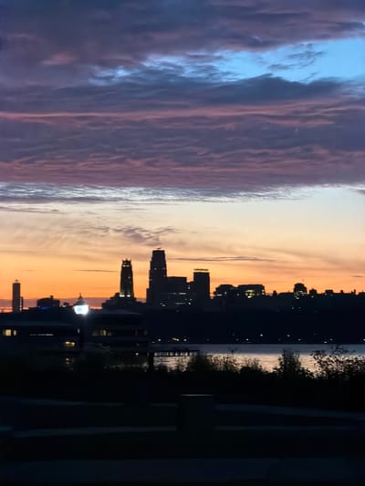 The image captures a city skyline at dusk, likely Edgewater, United States of America. The sky is painted with a gradient of colors, transitioning from deep blues and purples at the top to vibrant oranges and pinks near the horizon. Wispy clouds stretch across the sky, catching the fading light. Silhouetted against this dramatic backdrop are the distinct shapes of buildings, some tall and modern, others with more traditional architectural features. A body of water, possibly a river or bay, lies in the foreground, its surface reflecting the colors of the sky. The immediate foreground is dark, with the silhouettes of trees and vegetation, and a dimly lit path or walkway is visible. The scene evokes a sense of calm and transition as day gives way to night.