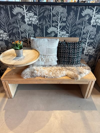 This interior scene, located in Altenheim, Germany, presents a light-colored wooden bench positioned against a wall covered in dark blue wallpaper featuring a white or silver pattern of stylized trees and foliage. The bench has a solid wooden seat and a slatted wooden backrest. On the bench, from left to right, a round wooden tray holds a small galvanized metal pot containing a green plant with numerous small orange flowers. To its right, an off-white decorative pillow with geometric patterns, fringe, and metallic embellishments rests against the backrest. Adjacent to it, a dark green and black woven tweed-like pillow is also positioned. A light brown faux fur rug or throw is draped across the bench seat, extending partially beneath the pillows. The floor is a light-colored, smooth surface, possibly concrete, exhibiting shadows cast by the bench and its contents. No individuals are present, and no actions are observed within the frame.