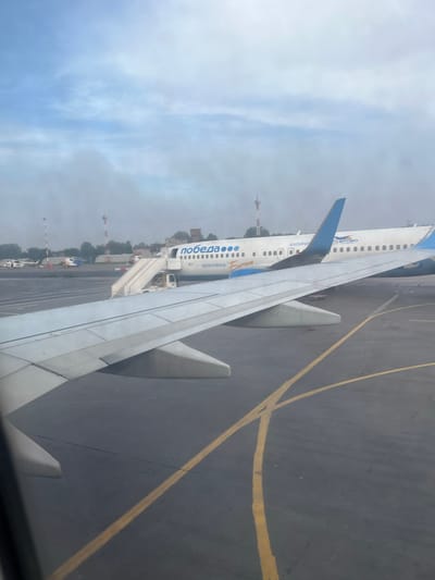 The image depicts an airport tarmac on a cloudy day, likely in or near the city of Sokol, Russia. In the foreground, the wing of an aircraft is visible, extending from the left side of the frame. Beyond the wing, a white passenger airplane with blue accents is parked. The plane has the word "ПОБЕДА" and "ЖЕМЧУЖИНА БАЛТИКИ" written on its fuselage. A mobile set of stairs is attached to the airplane's door, indicating passengers may be boarding or disembarking. In the background, other aircraft and airport buildings can be seen, along with radio towers. The tarmac is marked with yellow lines. The overall atmosphere is hazy, suggesting mist or low visibility. No people are clearly visible in the image.