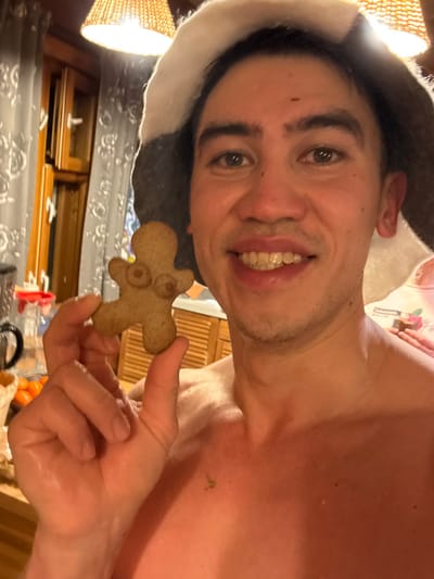 A smiling man wearing a sauna hat holds a gingerbread man cookie with two circular markings on its chest. The man's skin is shiny, possibly from sweat. The background includes a woven lampshade, a wooden cabinet, and a glimpse of a window. Other objects like a black container and oranges are partially visible on a surface below. The image has a slight upward angle.