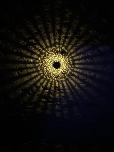 The image shows a close-up of a textured, dark wall illuminated by a light fixture. The light fixture, positioned at the center of the image, casts a patterned shadow that radiates outwards like a sunburst. The shadow pattern is intricate and appears to be made of decorative cutouts or a stencil. The wall itself has a rough, uneven surface, with visible indentations and variations in texture. The lighting creates a high contrast scene, with bright illuminated areas and deep shadows. The overall impression is one of ambient, decorative lighting on an aged surface. The location is specified as Villa Pasquali, Italy.