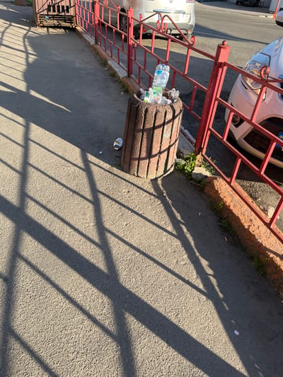 The image captures a street scene in Kalynivka, Ukraine, characterized by a concrete trash receptacle overflowing with plastic bottles and other debris. A red metal fence borders the sidewalk, casting distinct shadows from overhead structures onto the asphalt. Several cigarette butts are visible on the ground near the fence. In the background, cars are parked along the street, and a portion of a building or structure with a metal gate is also present. The lighting suggests it is daytime, likely late afternoon due to the long shadows.