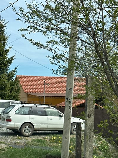 The image depicts a residential area in Raychikhinsk, Russia. A white station wagon is parked along a gravel driveway. A house with a red tile roof is visible behind the car, adjacent to a dark brown structure. Two utility poles stand in the foreground, partially obscured by tree branches with new spring leaves. Evergreen trees are also present in the scene. The sky is a clear blue with a few wisps of clouds. Written markings are visible on the utility poles.