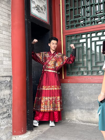 The image displays a young East Asian male posing in a doorway within a traditional Chinese architectural setting in Haidian District, China. The man, appearing in his late teens or early twenties, wears an elaborate, dark red traditional garment, possibly a Hanfu, richly embroidered with gold and colorful dragon and auspicious patterns. His attire includes a pleated lower skirt-like section and an upper robe with wide sleeves, cinched by a decorative white-buckled belt. Modern white and grey athletic sneakers are visible on his feet. He stands with both arms bent at the elbows, fists clenched, and a focused expression directed slightly left of the viewer.
To the left of the doorway, a smooth, cylindrical red pillar stands against an aged grey brick wall. Above the doorway, a framed traditional Chinese landscape painting, featuring mountains and water, is integrated into the wall. To the right of the doorway, a wall section contains two stacked windows, each fitted with intricate dark green wooden lattice patterns set within red frames. On the far right, the right arm and shoulder of a second person are partially visible. This individual holds a black smartphone, aimed at the posing man, indicating they are taking a photograph. The photographer is wearing a light-colored top and a teal athletic short or skirt. The ground is a light grey concrete surface, showing signs of wear.