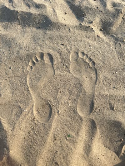 Two footprints are imprinted in the fine sand. The footprints are of a person's feet, showing the distinct shape of toes and the sole. The sand appears light brown and slightly damp, suggesting it might have been recently wet, perhaps by the tide or recent rain. The lighting suggests it is daytime, with a shadow cast from the left, indicating the sun is at an angle, possibly morning or late afternoon. The scene is a natural, outdoor setting, likely a beach or sandy area. There are no people, objects, text, or discernible emotions visible in the image. The context of Noyabrsk, Russia, implies a cold climate, so this sandy scene could be a seasonal or protected area.