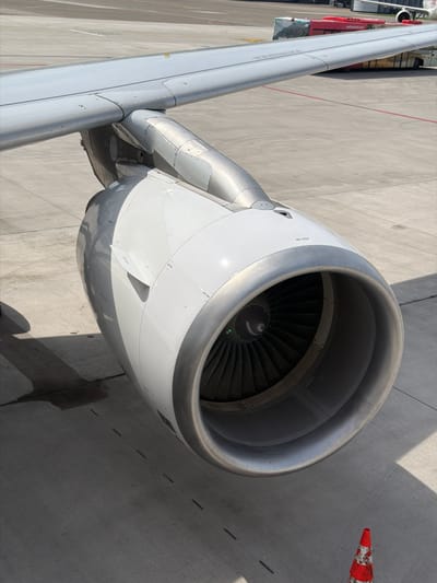 An overhead shot captures a large jet engine attached to an aircraft wing, both predominantly light gray and white, parked on a concrete airport tarmac. The sun casts strong, sharp shadows, suggesting bright daylight, possibly morning or late afternoon. The engine's intake, with its dark fan blades visible within, is a central focus.
Several details are noticeable:
- **Aircraft:** The smooth, metallic surface of the engine nacelle and the aircraft wing are visible. Markings such as "NO STEP" and "HOIST POINT" with an arrow are present on the engine cowling.
- **Ground Environment:** The concrete ground shows various seams and markings, including a red line partially visible on the left. A red and white striped safety cone lies on its side to the right of the engine, indicating an active apron area.
- **Ground Service Vehicle:** Part of a red ground service vehicle is visible near the wing root on the left side of the image, with "swissport.com" clearly printed on its side, confirming an airport operational context.
- **Engine Details:** The engine's fan blades are dark, and a small, bright green light or reflection is visible deep within the engine's center. The metallic lip of the engine intake reflects the sunlight.
The image portrays a typical scene at an airport, likely during ground operations, with the aircraft stationary. The absence of people emphasizes the machinery and infrastructure of air travel. The location context is Kloten, Switzerland, which is consistent with the presence of a Swissport vehicle, a common ground handling provider at Zurich Airport.