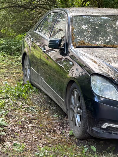 A dark-colored Mercedes-Benz is parked on a lawn covered in grass, leaves, and small weeds in Tokkari-Lend, Russia. The car is partially covered with debris such as leaves and pollen, suggesting it hasn't been moved in a while. The windows reflect nearby buildings. The car's front passenger side is visible, including the headlight, front tire, and side mirror.