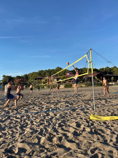 In Pistula, Montenegro, a group of young adults are engaged in a game of beach volleyball on a sandy court under a clear blue sky. The net, supported by yellow poles with the brand "COPAVA" visible, divides the court. One man is in mid-air, spiking the ball over the net, while another player on the opposing side watches. Several other players are positioned on the sand, anticipating the next move. In the background, lush green trees and a building with a thatched roof suggest a relaxed, coastal setting. A yellow bag with the words "Beach Volleyball" lies on the sand near the net.