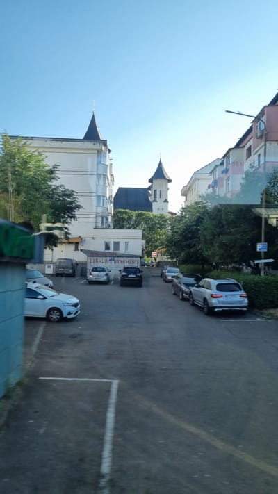 A street view in Fălticeni, Romania shows a paved parking lot with several cars parked. In the background, buildings line the street, including a white building with a distinctive turreted roof, and a taller white tower with a dark roof. A church is visible with a steeple. Trees are scattered throughout the scene. The sky is clear and blue. The ground in the foreground is asphalt with white parking lines. A blue object, possibly part of a vehicle or structure, is partially visible on the left side of the image. The text "BERMUR --ORNAMANTE" is visible on one of the buildings.