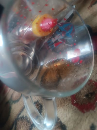 A clear glass mug is depicted from an overhead, angled perspective, showcasing its interior contents. The mug is adorned with scattered small red heart decals and a blue and red graphic illustrating two figures under an umbrella. Inside the mug, at the bottom, a brownish granular residue is present. Reflections on the glass surfaces partially obscure a yellow object with a pointed red tip and other indistinct metallic items within the mug's cavity. The mug rests on a patterned rug or carpet, which displays shades of brown and beige with subtle red designs. This scene is situated in the city of Elatan, Uzbekistan.
