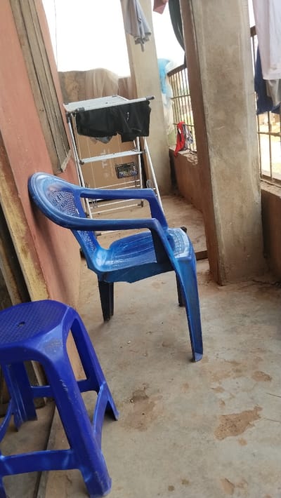 The image displays an outdoor or semi-outdoor domestic area, identified as a balcony or porch, located in Isieke, Nigeria. In the foreground, a blue plastic stool with a textured top surface rests on a light grey concrete floor. Adjacent to it, a blue plastic armchair with armrests leans against a reddish-brown wall or partition. To the right of the armchair, a partially collapsed silver-colored clothes drying rack is positioned. It holds two dark-colored garments over its upper bar, and a small black basket containing white items is on a lower shelf. A brown cardboard box, partially visible, is situated behind the drying rack. A vertical light grey concrete pillar divides the scene towards the right. Beyond this pillar, a metal security grate or railing is visible, adorned with various hanging laundry items, including white, grey, red, and blue fabrics. The background beyond the railing appears brightly lit, suggesting an open outdoor environment. The concrete floor exhibits multiple stains and discolorations. No individuals are present in the scene.