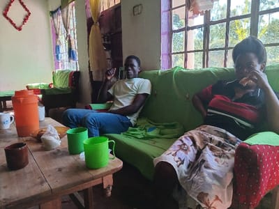 The image captures a casual indoor scene, likely a living room, in Manda-Shivanga ward, Kenya. Two individuals are seated on couches covered with green fabric.
On the left, a man is comfortably seated, wearing a light-colored (possibly cream or light yellow) t-shirt and blue jeans. His left hand is raised towards his chin, suggesting a moment of thought or relaxation. To his right, a woman sits on a separate couch. She is dressed in a red, black, and white striped shirt and a long, light-colored patterned skirt, possibly with floral designs. She appears to be eating a piece of food, holding it to her mouth with her right hand.
In the foreground, a wooden coffee table holds several items. These include a large orange thermos, two bright green plastic mugs, a brown ceramic mug, and a white ceramic mug. A small white bag or cloth, possibly containing food items like bread or snacks, is also on the table next to a light-colored bowl.
The room is illuminated by bright natural light streaming through a large window in the background, which reveals glimpses of lush greenery outside, suggesting a sunny daytime. The window is adorned with colorful, sheer curtains in shades of blue, purple, and yellow. A red, diamond-shaped decoration hangs on the wall to the left. The overall setting is simple and domestic. There is no discernible text visible in the image.