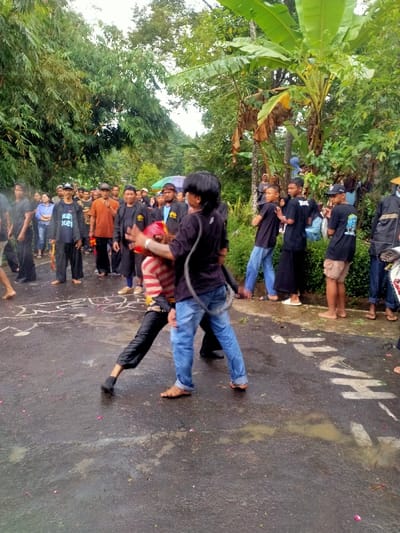 The image depicts an outdoor scene on a wet asphalt road in Mojogedang, Indonesia, bordered by dense green vegetation. In the foreground, two barefoot males are engaged in a dynamic interaction. The individual on the left wears a red and white striped long-sleeve shirt, dark pants, and a red head covering; they are in a low, athletic stance with the left leg extended forward and the right arm raised. The individual on the right, facing the first, wears a black short-sleeve shirt and blue jeans, with a dark coiled rope slung over their left shoulder; their right hand is raised with the palm open.
Approximately 20-30 individuals, predominantly males dressed in dark shirts and pants, are gathered in the midground along both sides of the road, observing the central figures. Some spectators are barefoot, while others wear footwear or are partially obscured. Scattered pink and red flower petals are visible on the wet road surface, along with white painted markings: an abstract, elongated shape to the left of the central figures and the partial word "HATI-" written in block letters to the right. A small puddle is also present on the road. The background consists of lush, tall green trees, including prominent banana plants, under an overcast sky.