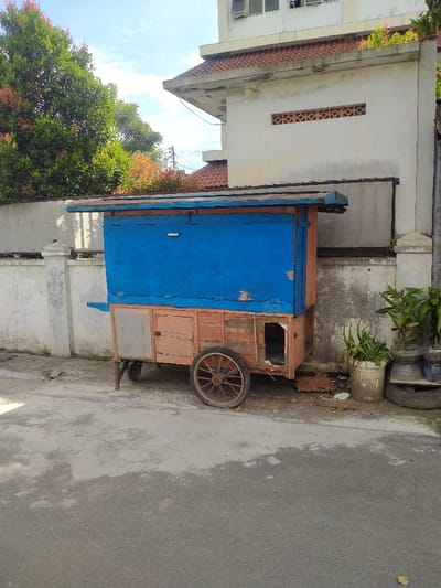 The image shows a blue and brown food cart parked on a paved street in front of a white building. The cart is made of wood and has a blue roof. The cart has one large wheel and two smaller wheels. The cart is parked on a street in front of a white building. The building has a red tiled roof. There are trees and plants in the background. The location is in Kecamatan Kartasura, Indonesia. The coordinates are latitude: -7.566, longitude: 110.776. The image was taken on a sunny day. The sky is clear. The street is empty. There are no people in the image. The cart is empty. The building is empty. The trees are bare. There is a small pile of garbage in the foreground. The image appears to be a still shot. The image has a shallow depth of field. The cart is in focus. The building and the background are out of focus.