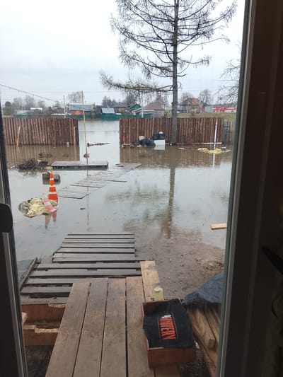 The image shows a flooded area, presumably in Высокиничи, Russia, viewed from inside a building, possibly through an open doorway. The water level is high, covering the ground and reaching up to a wooden fence in the background. Debris and various objects, including what appears to be insulation material and an orange traffic cone, are floating in the water. Makeshift wooden walkways are visible, possibly to navigate the flooded area. A bare tree is visible in the mid-ground. Inside the doorway, there is construction or storage of wooden planks and an object with the word "Mystery" written on it in English. The weather appears overcast and rainy.