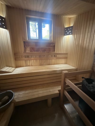 The image shows the interior of a wooden sauna. The walls and ceiling are constructed of light-colored wooden planks. There are two levels of wooden benches, with the upper bench being narrower. A small window with two panes is visible above the upper bench, offering a view of trees and a bright sky. Two square wall-mounted lights with multiple small bulbs are present, one on the left and one on the right side of the room. A small wooden stool is placed on the lower bench on the left side of the image. A metal bucket with a wooden handle is on the floor to the left. In the foreground on the right, part of a wooden structure, possibly another bench or a support, is visible, along with a black object that appears to be a sauna heater with stones and a ladle. The overall impression is of a clean and functional sauna space. The location is Brca, Montenegro.
