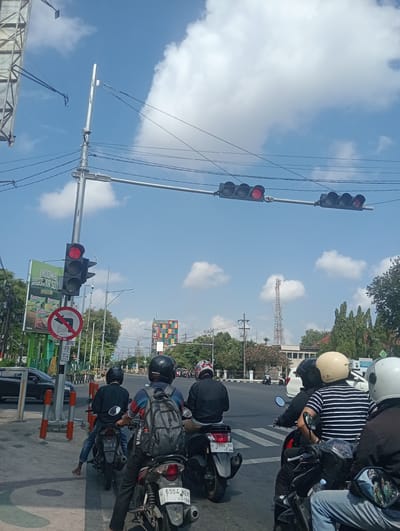 The image depicts a street intersection in Sidoarjo, East Java, under a bright, partly cloudy sky. A traffic light is prominently displayed, with the red light illuminated on the left-hand signal and an arrow pointing left illuminated on the right-hand signal. Several motorcycles are stopped at the intersection, indicating a traffic pause. Riders are wearing helmets, and some are carrying backpacks. A billboard advertising "Indomie Jagung" is visible to the left, along with a traffic sign indicating no left turn. In the background, there are trees, buildings, and a telecommunications tower. The overall scene suggests a typical urban traffic scenario in Sidoarjo.