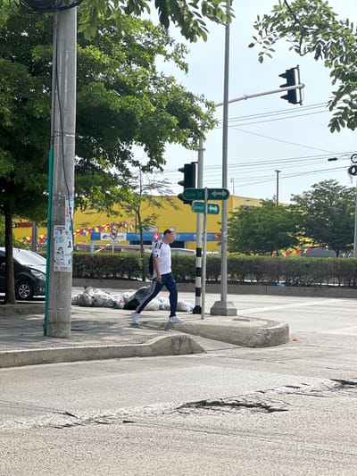 A man wearing a white t-shirt with black stripes on the sleeves, dark pants with white stripes, and white sneakers is walking on a sidewalk next to a street in Perímetro Urbano Barranquilla, Colombia. He is carrying a black backpack and appears to be walking towards the right of the frame. In front of him, on the sidewalk, are several large white garbage bags. To his left, a light pole with various signs and wires is visible, as well as a dark-colored car parked on the street. Across the street, a long, yellow building with colorful banners is in the background, bordered by a line of green bushes. Traffic lights and street signs indicating "Calle 30" and "Carrera 8" are also present. The road surface shows signs of wear and tear, with some cracked areas. The sky is bright and overcast, suggesting daytime.