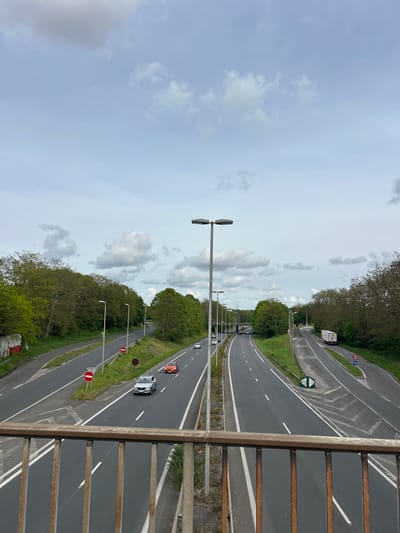 A vertical shot from a bridge looking down at a highway. Two three-lane highways run parallel to each other, separated by a grassy median with trees. Street lights line the median and the outside of each highway. Vehicles are visible on both highways, including cars and a truck. The sky is overcast with clouds. A rusted metal railing is visible in the foreground of the bridge. The highway is in Noisiel, France.