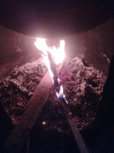 The scene, located in Nurli zamin, Uzbekistan, depicts an active fire contained within a dark, enclosed structure. Bright, reddish-orange flames, featuring intermittent blue and purple hues, originate from burning wooden fuel situated centrally within the frame. A partially consumed wooden plank is visible, extending diagonally from the lower-left towards the center-right, contributing to the combustion. Below and surrounding the flames, a significant accumulation of glowing embers and greyish-white ash occupies the lower portion of the enclosure. The surrounding environment appears dark and textured, consistent with the interior of a stove or fire pit, with a dark, curved overhead surface. No individuals, specific actions, or additional objects are present.