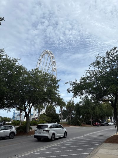 This image depicts a street scene in Panama City Beach, Florida. In the foreground, a white minivan is driving on a paved road. To the left, a silver SUV is parked. Several large, leafy green trees line the side of the road, with some smaller palm trees interspersed. In the background, a large white Ferris wheel is visible against a cloudy sky. The Ferris wheel appears to be part of an amusement park or entertainment venue. The overall environment suggests a coastal city with a tourist-oriented atmosphere. The sky is partly cloudy with a pattern of elongated clouds. There are no people clearly visible in the image.