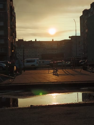 The image depicts a sunset over an urban landscape in Norilsk, Russia. The sun, a bright orb, is low on the horizon, casting a warm, golden light across the scene. Silhouettes of apartment buildings dominate the background, their windows reflecting the fading sunlight. In the foreground, a paved area with a puddle of water reflects the sky and the surrounding structures. Cars are parked along the street, their forms softened by the low light. A lamppost stands tall, its shadow stretching across the pavement. The overall atmosphere is one of a quiet, end-of-day stillness in a northern city.
