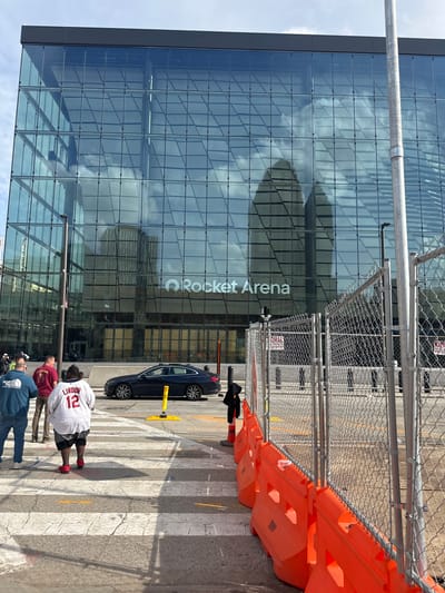 The image depicts Rocket Mortgage FieldHouse in Cleveland, Ohio. The building's glass facade reflects the sky and surrounding structures. Construction barriers and fencing are present. A black car is parked near the entrance. Pedestrians, including one wearing a "Lindor 12" jersey, are walking across a crosswalk in front of the arena.
