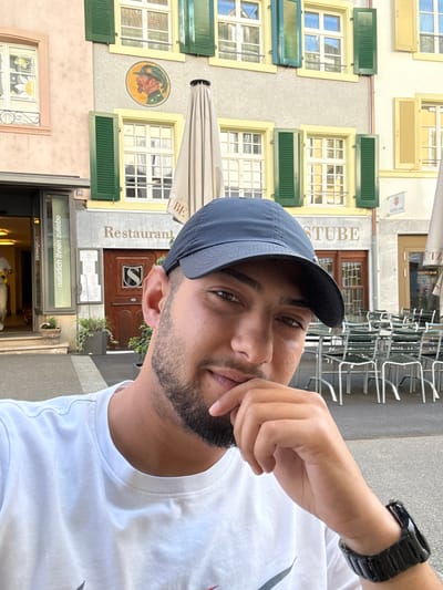 The image is a daytime selfie of a man outdoors, framed from the chest up. He has dark hair, a beard, and brown eyes, looking directly at the camera with a neutral to slightly contemplative expression. He wears a dark blue baseball cap and a white t-shirt, with a black-banded watch on his left wrist. His right hand rests near his mouth and chin.
The background reveals a traditional European building, likely a restaurant or inn, set in an urban environment consistent with Liestal, Switzerland. The multi-story structure features light-colored walls with numerous windows framed in yellow, many adorned with green shutters, some open and some closed. A round, colorful mural depicting a person's head in profile is painted on the building's facade.
Several signs are visible on the building. The prominent text "Restaurant" and "STUBE" identify the establishment. Below the main entrance, a glass panel reads "natürlich ihnen zuliebe," which is German for "naturally for your sake" or "naturally to please you." Above the "STUBE" sign, another sign states "SCHÜTZEN BAR." The number "16" is marked near a doorway. To the right, an outdoor seating area with numerous stacked metal chairs suggests a patio or café setting. The bright, clear lighting indicates it is daytime with pleasant weather.