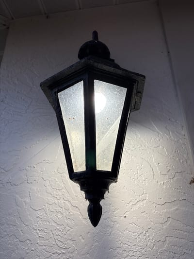 A black, ornate lantern hangs on a textured white wall. The lantern has clear glass panels with a visible light bulb inside that is illuminated, casting a bright glow. The wall has a rough, stucco-like finish with visible indentations and ridges. Above the lantern, a white ceiling or overhang is visible, featuring a grid pattern. The lighting suggests it might be dusk or a dimly lit area. The image is taken from a low angle, looking up at the lantern.