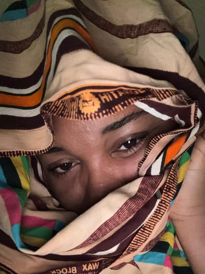 A close-up shot shows a person's face partially covered by a patterned fabric. Only the eyes, forehead, and a portion of the nose are visible. The fabric has a complex design with stripes in brown, beige, orange, white, and other colors. Part of the fabric has the text "XAW BROCK" printed on it. The scene is set in Rumuokwuta, Nigeria.