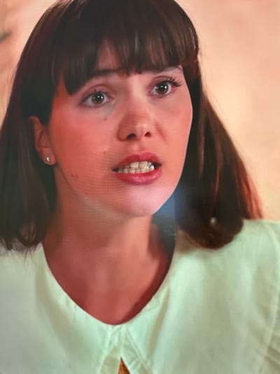 The image shows a close-up of a young woman's face and upper chest. She has dark brown hair with bangs, brown eyes, and full lips with red lipstick. She is wearing a light green collared shirt. She has a small stud earring in her left ear. The background is a soft, peachy-orange color. There is no indication of the city of Uyutnoye, Ukraine in the image.