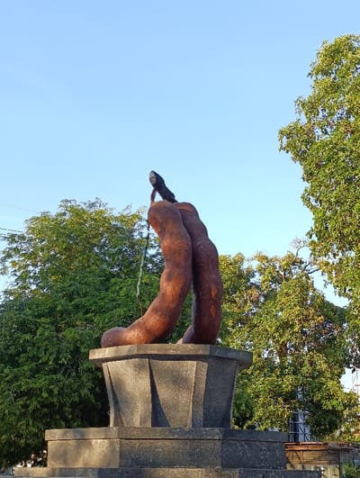 The image shows a sculpture of two intertwined, brown, abstract shapes on top of a square, grey, stone base. The sculpture is located in a park in Kecamatan Asembagus, Indonesia. The base is made of stone and has a square base with four sides. The sculpture is surrounded by green trees and a clear, blue sky. The sculpture is located on a pedestal that is visible in the lower half of the image. There are no people or other subjects in the image. The sculpture appears to be made of a smooth, brown material. There are no visible markings or details on the sculpture. The sky is clear and blue, and the sun is shining. The trees are green and leafy, and the leaves are casting shadows on the ground. The grass is green and lush. The image appears to have been taken during the day, as the sunlight is casting shadows.