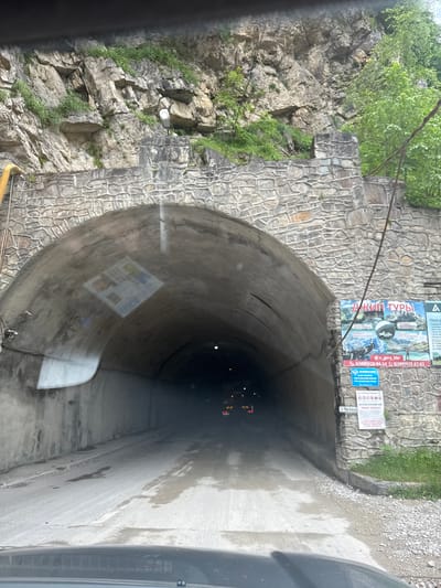This media file captures a view from inside a vehicle as it enters a tunnel carved into a rocky mountainside. The tunnel's interior is dark and appears damp, with evidence of water on the walls and road. The entrance is framed by a rough stone wall, and a cliff face covered in greenery is visible to the left, suggesting a mountainous or rugged natural setting.

Several signs are posted on the stone wall near the tunnel entrance. One prominent sign, with Cyrillic text, appears to be advertising "ДЖИП ТУРЫ" (Jeep Tours). Other smaller signs are also visible, including what looks like a price list or informational material, and a blue sign with Cyrillic text. A license plate reading "C964KA" is also visible near the tunnel opening.

The overall impression is a transition from an outdoor, natural environment into a man-made structure. The lighting suggests it might be daytime, but the tunnel's interior is dim, with faint lights visible further inside, possibly from other vehicles. The scene is devoid of people, focusing on the environment and the infrastructure. The image conveys a sense of journeying into a more enclosed and potentially adventurous space, indicated by the "Jeep Tours" advertisement.