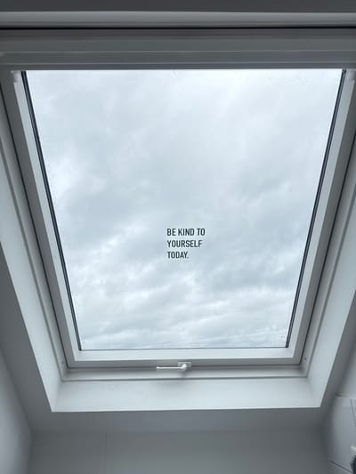 The image depicts a white-framed window with a view of a cloudy sky. The window pane has the words "BE KIND TO YOURSELF TODAY." printed on it. The window is located in Ampfing, Germany. The room is otherwise plain and white. No people or interactions are visible.