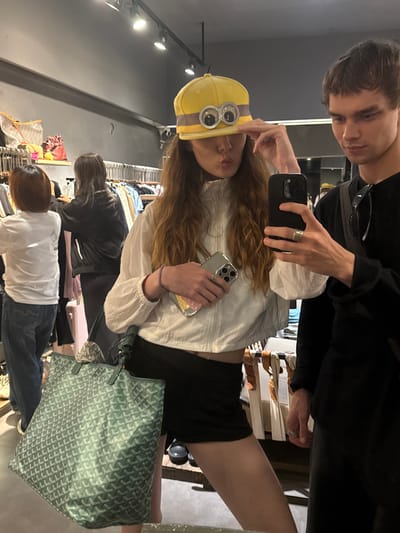 In Xuhui District, China, a young woman wearing a yellow hat with googly eyes and a white jacket is taking a selfie with a young man in a black t-shirt. The woman is holding a phone in her left hand and is making a pouting face. She is also wearing a short black skirt and carrying a green tote bag. The man is wearing glasses and has his arm around the woman. In the background, there are racks of clothing and bags, suggesting that they are in a store.