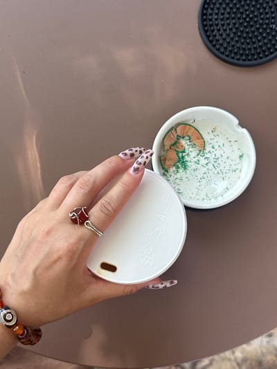 The image provides an overhead view of a person's right hand and various objects arranged on a smooth, brown table surface, set in Kaş, Turkey.

The primary subject is a person's hand, adorned with long, coffin-shaped nails featuring a distinctive black and white spotted pattern, resembling leopard print. A silver wire-wrapped ring with a prominent dark red stone, possibly carnelian or garnet, is worn on the middle finger. A beaded bracelet, showcasing various shades of orange, brown, and black, along with a silver accent bead, is visible on the wrist.

The hand is holding a white circular object, likely a cup lid, which has a small brown-rimmed opening for drinking. Faint, stylized symbols or text, possibly in an East Asian script, are subtly embossed or printed on the surface of the lid.

To the left of the hand, a white, circular ceramic object, resembling an ashtray, contains several curled, light brown pencil shavings and numerous small, bright green fragments, suggesting it has been used to collect pencil sharpenings. Further to the upper left, a black, circular rubber coaster with a textured, studded surface is also visible on the table. The background features a blurred, lighter-toned, textured surface, possibly a floor or another table. The scene captures a casual moment, potentially indoors or in a shaded outdoor area, given the ambient lighting and reflections on the table.