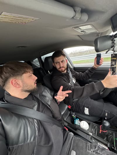 Inside a moving vehicle in Gabarnitsa, Bulgaria, two young adult males are visible. The scene is captured from the rear passenger side, facing forward.

In the front passenger seat, a male with dark hair, a short beard, and a black puffer jacket with a visible seatbelt is turned towards the driver. His mouth is slightly open, suggesting speech or reaction.

The driver, positioned in the left foreground of the frame, is a male with dark hair and a full beard, wearing a black hooded sweatshirt and dark pants. He holds a dark blue smartphone in his right hand, pointed forward and slightly to the right, while his left hand is raised in a gestural pose towards the passenger. His expression indicates engagement, possibly a strong point or reaction. A black rosary-style chain with a crucifix and an "areon" branded air freshener hangs from the rearview mirror.

The car's interior features a dark gray dashboard and upholstery, a light gray headliner, and a center console containing a black Monster energy drink can, a light blue cigarette pack, and a white disposable coffee cup. A gear shift with a black knob wrapped in a red braided cord is also present. Through the front and side windows, a multi-lane road with a guardrail structure and an overcast sky are visible.