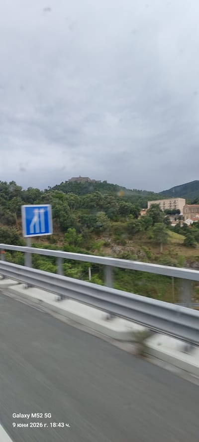 A cloudy day scene captured from a moving vehicle shows a highway guardrail in the foreground, with a blue traffic sign featuring two white arrows indicating a recommended lane change to the left. Beyond the guardrail, a lush green hillside is covered with trees, and on its summit, a historical fortress or citadel is visible. Further in the background, a large, modern residential building and a section of an older building with red-tiled roofs are present. The image also contains a timestamp in the lower left corner: "Galaxy M52 5G 9 юни 2026 г. 18:43 ч.". Based on the presence of the citadel and the landscape, this scene is likely located in the vicinity of Le Perthus, France, a town known for its historical fortifications.