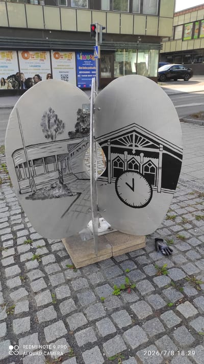 The image shows an outdoor public art installation on a cobblestone street in Iisalmi, Finland. The artwork consists of two large, silver, oval-shaped panels joined by a metal support. Each panel features a black and white drawing. The left panel depicts a building with a covered porch and trees. The right panel shows an architectural facade with arched windows and a large clock face with one hand pointing to approximately 7 o'clock. A black glove lies on the cobblestones to the right of the artwork. In the background, a traffic light is visible, as are posters for events, including "Oluset" and "LIMUSIN." A dark-colored car is parked on the street in the background. The timestamp in the lower right corner indicates July 8, 2026, at 18:29.