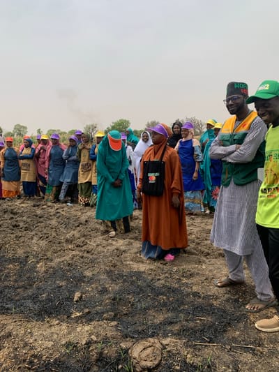 A group of approximately 20 people stand on a field of dark, ashy soil in Ringimawa, Nigeria, at coordinates 11.667 latitude and 8.416 longitude. There are a few trees in the background. The people are mostly women wearing colorful hijabs and long dresses. Two men stand on the right side of the image; one is wearing a green vest and striped shirt, while the other is wearing a green vest and a green shirt with a logo printed on it. Most of the women look at the camera with serious expressions on their faces. The men are looking to their right.
