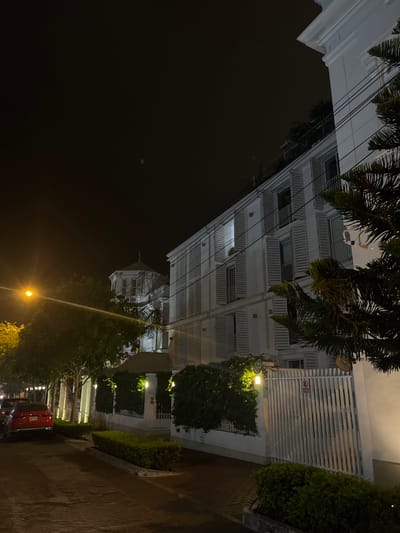The image shows a street scene in Barranco, Peru at night. A tall, multi-story white building with multiple windows, balconies, and a domed structure is visible on the left side of the image. A gate is present with horizontal slats. The right side shows vegetation. Streetlights illuminate the area, casting a warm glow. On the street, multiple parked cars are visible, including a red one. A sidewalk with a curb runs along the right side. The sky is dark. The image is rotated counter-clockwise.