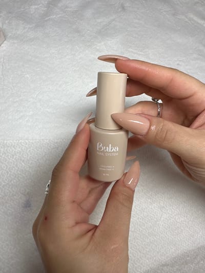 The image displays a close-up view of two hands holding a bottle of nail product against a white, textured background, likely within the city of Kafr Kasim, Israel. The hands are manicured with long, pointed nails coated in a light beige polish. The right hand wears a silver-colored ring with a clear stone on the ring finger, and the left hand has a plain silver-colored ring on the pinky finger. A small red mark or scab is visible on the back of the left hand near the thumb. The product bottle is light beige with a matching cap, labeled "Buba NAIL SYSTEM." Below the brand name, the label specifies "TPO FREE X" and "HEMA FREE X," followed by "16 ML" at the bottom. The hands are positioned to showcase the bottle, holding it upright. A blurry pink object is partially visible in the upper left corner of the background.