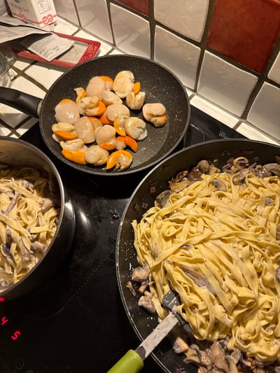 Three pans sit on a black stovetop, one containing sautéed scallops with orange roe, and the other two holding fettuccine with mushrooms and creamy sauce. A green-handled spatula is partially visible in the larger pasta pan. The stove sits adjacent to a tiled wall in red, white, and gray. A box of ingredients sits just off the frame.